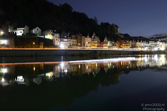 A nighttime view of a riverside town with illuminated buildings and a bridge reflecting in the - image from year 2026 #006