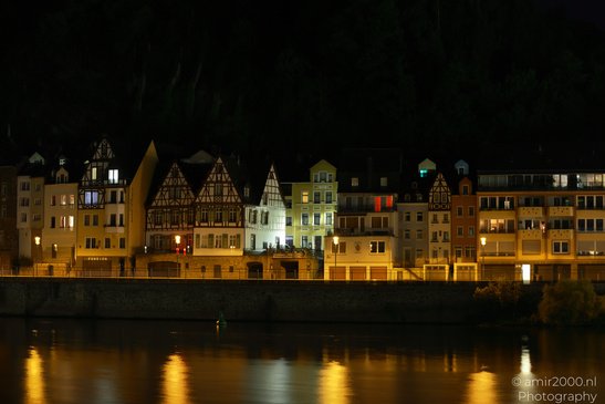 A row of illuminated half timbered houses along a riverbank at night, with reflections of the - image from year 2026 #010