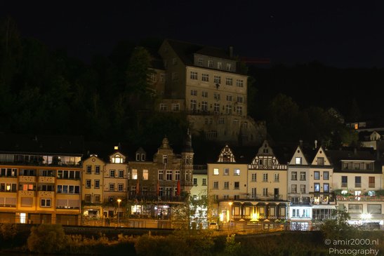 A row of illuminated buildings along a riverside at night, with a larger building perched atop a - image from year 2026 #015