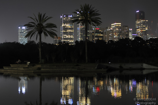 A nighttime cityscape with illuminated skyscrapers reflected on calm water, palm trees in the - image from year 2026 #001